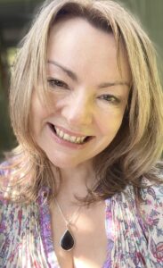 Women with sandy blonde hair and dark tips, smiling at camera in selfie. Wearing a necklace and multicolor soft floral top.