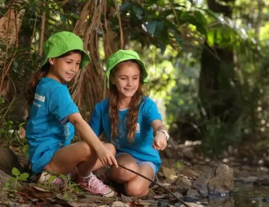 Two girls outside holding sticks and facing them towards pebbles on the ground