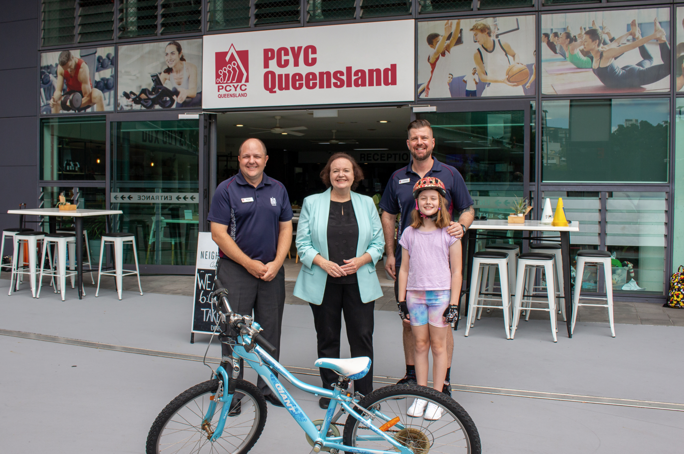 Staff, government and child posing in front of a PCYC Club with a bike in frame