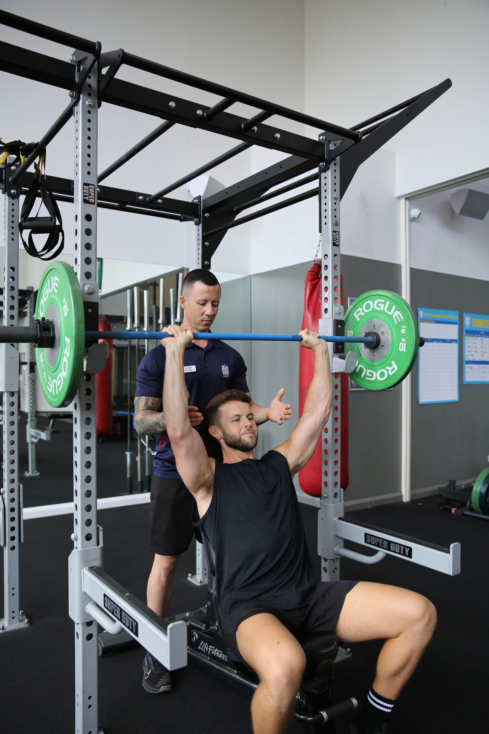 man performing lift at gym and being spotted by staff
